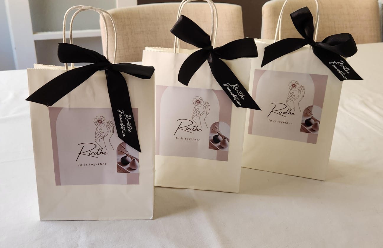 Three cream Rirothe Foundation gift bags with black ribbons and the "In it together" label, side by side on a table.