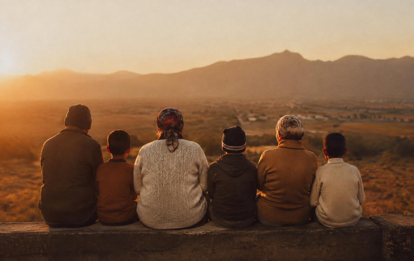 People of different ages sitting together on a low wall, looking out over a valley at sunset.